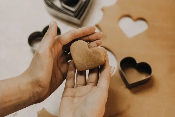 Mâini modelând o turtă dulce în formă de inimă, lângă forme de decupat în formă de inimă